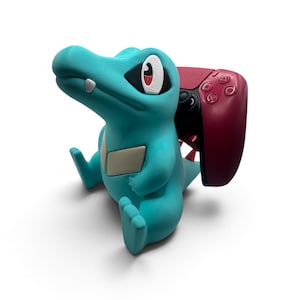 May include: A turquoise, crocodile-like creature figurine with a cream-colored chest plate, large eyes, and a single visible tooth, serving as a stand for a dark red video game controller that is positioned behind the figure's back.