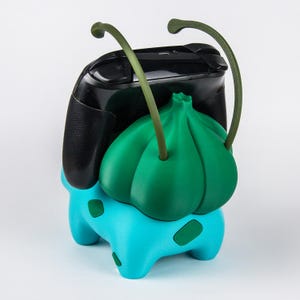 May include: A turquoise and green gaming controller stand shaped like a creature with a bulbous plant-like back and two long, curved antennae. The stand features dark green spots on its turquoise body and holds a black gaming controller.