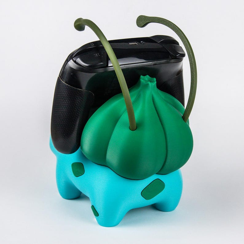 May include: A turquoise and green gaming controller stand shaped like a creature with a bulbous plant-like back and two long, curved antennae. The stand features dark green spots on its turquoise body and holds a black gaming controller.