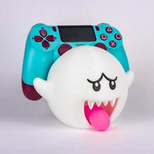 Può includere: Una figura di fantasma bianco con una lingua rosa e denti affilati, abbinata a un controller di gioco turchese e bordeaux. Il controller ha pulsanti neri e un touchscreen. Il fantasma ha sopracciglia nere e la bocca spalancata.