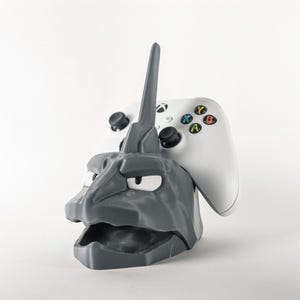 May include: A gray, 3D-printed controller stand shaped like a stylized monster head, holding a white Xbox controller. The stand has a horn-like protrusion and a wide open mouth. The controller has colored buttons.