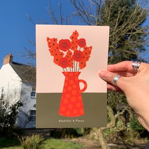 Framed wall art print in red, orange and olive green featuring gladioluses and the poppies in a polka dot vase. There is text below the vase in a hand-written font that lists the flowers.