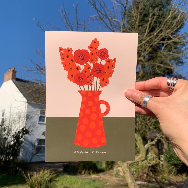 Framed wall art print in red, orange and olive green featuring gladioluses and the poppies in a polka dot vase. There is text below the vase in a hand-written font that lists the flowers.