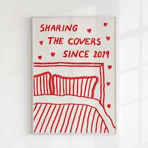 May include: A white framed print with red text that reads "Sharing the Covers Since 2019". The print features a red illustration of a bed with two pillows and a blanket.