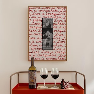 May include: A framed artwork with the words "Love is everywhere" in red script, and three black and white photos. A wine bottle, two wine glasses with red wine, and a bunch of grapes sit on a red serving cart.