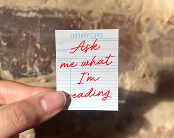 May include: A small white library card with blue lines and the words "Ask me what I'm reading" in red script. The card is held by a hand against a brick background. The words "Library Card" are printed at the top.