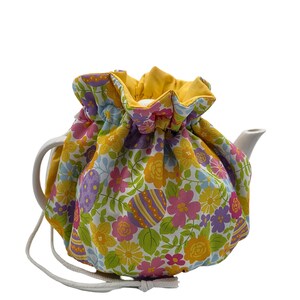 May include: A white teapot with a yellow and white floral print tea cozy. The cozy has a drawstring closure and is decorated with colorful Easter eggs.