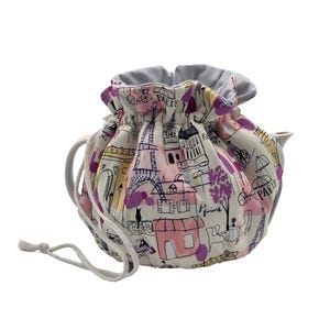 May include: A white teapot cozy with a drawstring closure. The cozy is decorated with a pink, purple, and yellow Parisian-themed print featuring buildings, a bicycle, and the word "Paris".