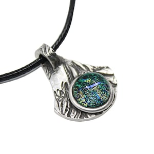 May include: A silver-toned pendant necklace with a round, dichroic glass stone in shades of green and blue. The pendant has a textured, organic design and hangs from a black cord. The necklace is a unique piece of jewelry.