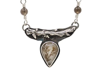 May include: A silver necklace with a pendant. The pendant has a teardrop-shaped, clear stone with a brown, tree-like inclusion. The pendant hangs from a silver chain with a branch-like design and two smoky quartz beads.