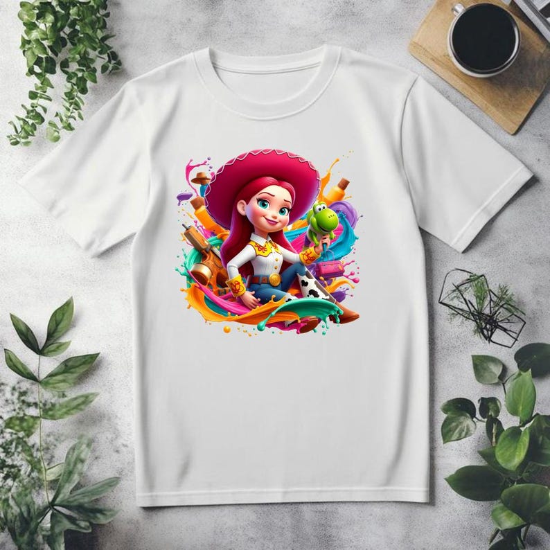 May include: White t-shirt featuring a colorful graphic of a cartoon cowgirl with a pink hat, holding a green toy dinosaur. The design includes paint splashes in various colors. The shirt has a crew neck and short sleeves.