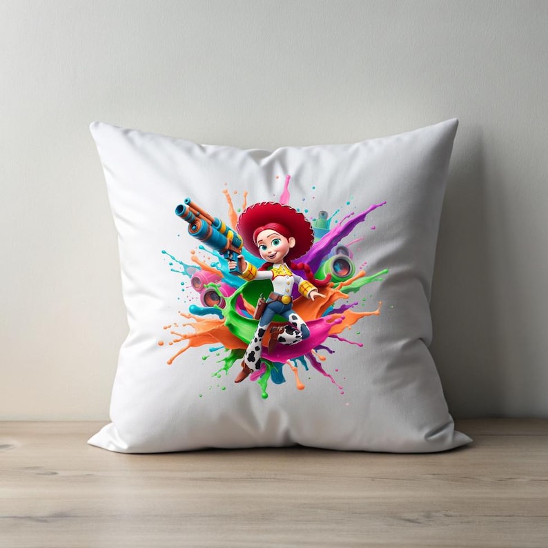 May include: White square pillow featuring a colorful illustration of a cartoon cowgirl holding a toy gun. The cowgirl is surrounded by a vibrant splash of paint in various colors, including orange, green, pink, and purple. The pillow is on a wooden surface.