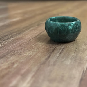 Puede incluir: Un anillo pequeño y redondo con una superficie facetada en color verde azulado intenso. El anillo se encuentra sobre un fondo de madera con un patrón de vetas natural, creando un contraste de texturas y colores. El anillo parece ser de metal.