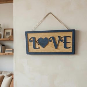 Puede incluir: Letrero de madera con la palabra "LOVE" en letras azul marino y un corazón. El fondo es de madera clara, enmarcado en azul marino y colgado de una cuerda. Un elemento decorativo para la decoración de interiores.