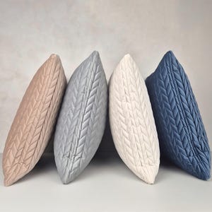 Peut inclure: Quatre coussins décoratifs avec un motif matelassé tressé. Les coussins sont dans des tons beige, gris, blanc et bleu.