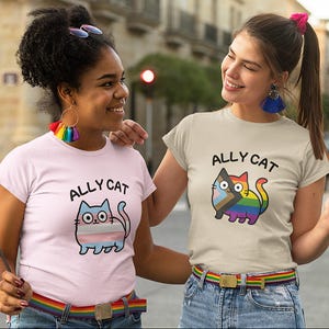 Könnte beinhalten: Zwei Personen tragen T-Shirts mit einem Cartoon-Katzen-Design und dem Text "ALLY CAT". Die Katzenillustrationen zeigen Regenbogen- und Transgender-Pride-Flaggenfarben. Beide Personen tragen Regenbogen-Gürtel.
