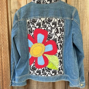 May include: A denim jacket with a large, colorful flower applique on the back. The flower has red petals, blue accents, and a yellow center. The collar features a black and white pattern. The jacket is hanging on a wooden surface.