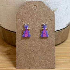May include: A pair of purple dragon stud earrings with pink accents. The earrings are on a brown card.
