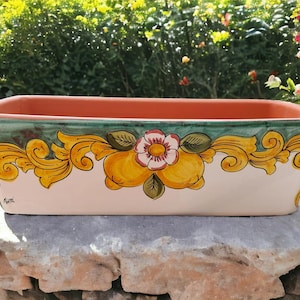 Può includere: Un vaso rettangolare in ceramica con fondo bianco e un disegno floreale verde, giallo e rosa. Il vaso ha un bordo leggermente arrotondato e un bordo in terracotta.
