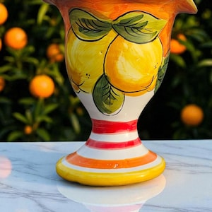 Puede incluir: Un jarrón de cerámica pintado a mano con borde acampanado y base de pedestal. El jarrón presenta un diseño vibrante de limones amarillos con hojas verdes sobre un fondo blanco. La base tiene rayas naranjas y rojas.