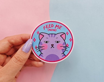 May include: A blue and pink round sticker with a purple cat with pink cheeks and the text "FEED ME (NOW)" in pink.