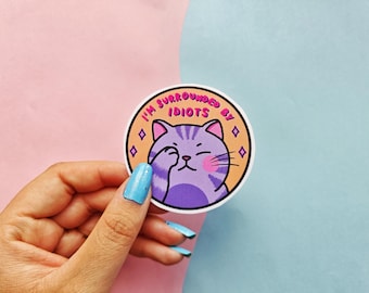 May include: A circular sticker with a purple cat covering its eye. The text "I'M SURROUNDED BY IDIOTS" is in a circle around the cat. The background is split between pink and blue.