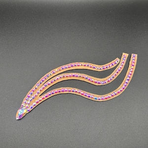 Op de afbeelding: Drie golvende, bruinachtige decoratieve applicaties, elk versierd met iriserende strass-steentjes. De strass-steentjes schitteren met een spectrum aan kleuren, waaronder roze, paars en blauw. De applicaties zijn in een gelaagd ontwerp gerangschikt.