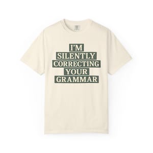 May include: A cream-coloured t-shirt featuring the text "I'M SILENTLY CORRECTING YOUR GRAMMAR" in dark green block letters. The shirt has short sleeves and a crew neck. The text is centred on the front of the shirt.