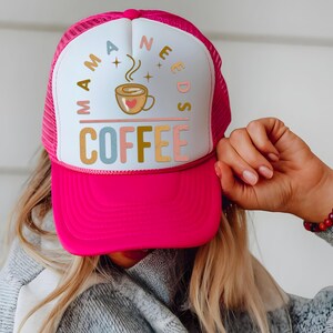 Puede incluir: Gorra de camionero rosa intenso con un panel frontal blanco. La gorra presenta el texto "MAMA NEEDS COFFEE" en varios colores, con un gr&aacute;fico de taza de caf&eacute;. La gorra la sostiene una persona que lleva un su&eacute;ter gris y una pulsera de cuentas.