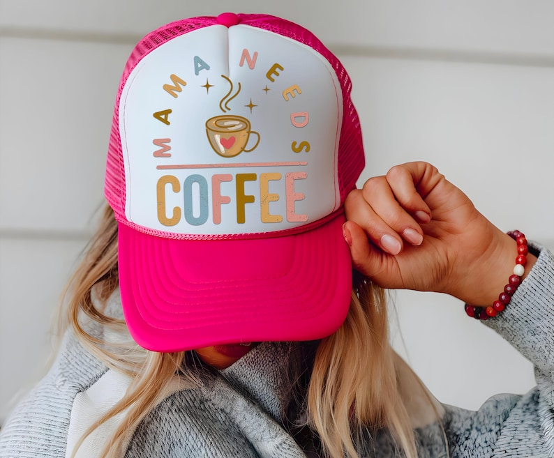 Puede incluir: Gorra de camionero rosa intenso con un panel frontal blanco. La gorra presenta el texto "MAMA NEEDS COFFEE" en varios colores, con un gr&aacute;fico de taza de caf&eacute;. La gorra la sostiene una persona que lleva un su&eacute;ter gris y una pulsera de cuentas.