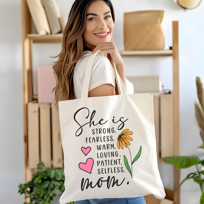 Puede incluir: Bolso tote de color crema con las palabras "She is" en fuente cursiva, seguido de palabras descriptivas y "mam&aacute;". Una flor amarilla y dos corazones rosas tambi&eacute;n est&aacute;n en la bolsa. La bolsa es sostenida por una persona.