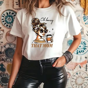 Puede incluir: Camiseta blanca con un gr&aacute;fico de una mujer con gafas de sol y una diadema con estampado de leopardo, sosteniendo una taza de caf&eacute;. El texto de la camiseta dice "Oh honey I am THAT MOM" en negro y dorado.