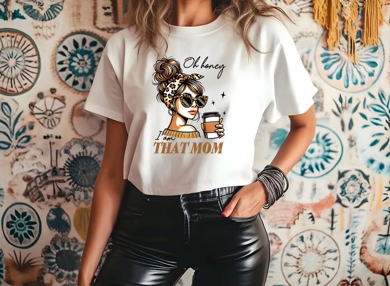 Puede incluir: Camiseta blanca con un gr&aacute;fico de una mujer con gafas de sol y una diadema con estampado de leopardo, sosteniendo una taza de caf&eacute;. El texto de la camiseta dice "Oh honey I am THAT MOM" en negro y dorado.