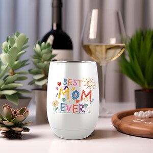 Puede incluir: Vaso de vino sin tallo blanco con las palabras "BEST MOM EVER" en letras coloridas de estilo manuscrito. El dise&ntilde;o incluye corazones, flores y un sol. Una botella de vino y una copa de vino est&aacute;n en el fondo.
