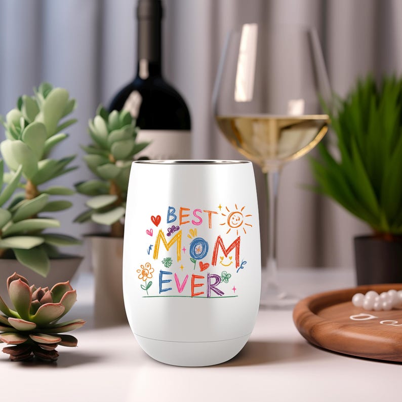 Puede incluir: Vaso de vino sin tallo blanco con las palabras "BEST MOM EVER" en letras coloridas de estilo manuscrito. El dise&ntilde;o incluye corazones, flores y un sol. Una botella de vino y una copa de vino est&aacute;n en el fondo.
