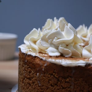 K&ouml;nnte beinhalten: Nahaufnahme eines runden Kuchens mit brauner Kruste und einer Schicht wei&szlig;er Schlagsahne obenauf. Der Kuchen befindet sich auf einer wei&szlig;en Papierunterlage.