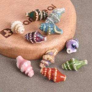 May include: A collection of ceramic seashell beads in various colors, including white, pink, blue, green, and red. The beads are arranged on a wooden surface and are ideal for jewelry making or craft projects.