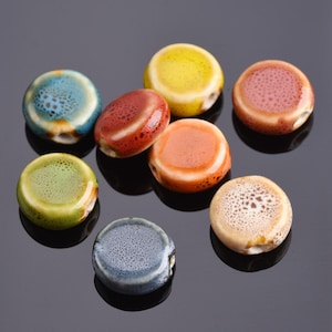 May include: A selection of colourful ceramic beads in shades of blue, green, yellow, orange, red, and tan. Each bead is round and flat with a hole for stringing. The beads have a speckled or mottled surface.