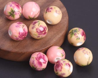 May include: A collection of ceramic beads in various shades of pink and cream. Each bead is adorned with a floral design in shades of pink and purple, with green leaves. The beads are round and have a hole for stringing.