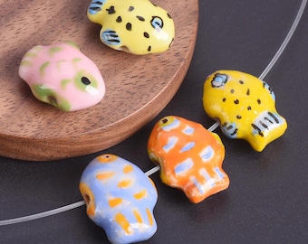 May include: A collection of colorful ceramic fish beads. The beads are in various colors, including yellow, pink, orange, and blue, with black dot and stripe details. They are strung on a clear cord, suggesting they are for jewelry making.