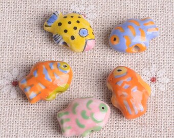 May include: Five colorful ceramic fish figurines. The fish are painted in bright colors including yellow, orange, blue, and pink. Each fish has a unique pattern and design, with details like spots and stripes. These small figurines are ideal for crafting or decorative purposes.