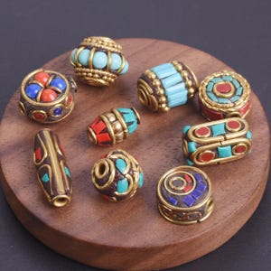 Pode incluir: Uma coleção de contas ornamentadas em várias formas e tamanhos, com elementos metálicos dourados e pedras incrustadas turquesa, vermelhas e azuis. As contas são exibidas em uma superfície redonda de madeira, mostrando seus intrincados designs e cores vibrantes.