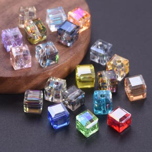 May include: A selection of multi-coloured, cube-shaped beads. The transparent beads reflect light, displaying colours such as purple, blue, green, yellow, and red. These beads are likely used for jewellery making or crafting projects.