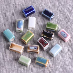 以下が含まれることがあります： 青、緑、白、茶色など、さまざまな色の長方形のセラミックビーズのコレクション。各ビーズは光沢のある仕上げで、紐を通すための穴があります。ビーズは、質感のある灰色の表面に散らばっています。