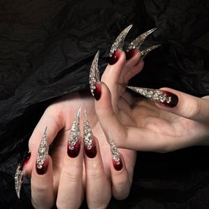 Puede incluir: U&ntilde;as artificiales largas y puntiagudas con un degradado de rojo oscuro a transparente, muy adornadas con pedrer&iacute;a plateada. Las u&ntilde;as son un accesorio dram&aacute;tico y glamuroso, perfecto para una ocasi&oacute;n especial o una declaraci&oacute;n de moda audaz.