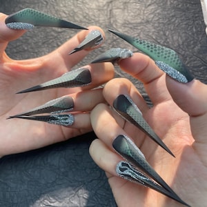May include: A set of long, pointed artificial nails. The nails feature a gradient design, transitioning from a dark green base to a black tip. The base of each nail is adorned with silver glitter, creating a striking contrast.