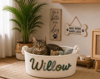 May include: A round, off-white woven cotton rope basket featuring the name Willow in dark green and grey cursive lettering on the front. The basket has two sturdy rope handles and contains a tabby cat along with several small plush pet toys.
