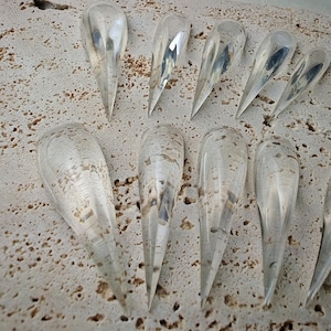 May include: A selection of clear, teardrop-shaped glass pendants with pointed ends. The pendants are displayed on a textured, light-coloured surface. The glass reflects light, creating highlights and a sense of depth.