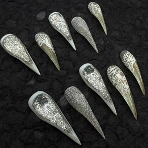 May include: A collection of silver and white artificial nails with a pointed shape. Some nails feature glitter, while others have a lace-like pattern. The nails are arranged on a textured black surface.