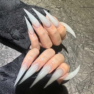 Puede incluir: Uñas acrílicas blancas largas y puntiagudas con purpurina. Las uñas tienen un efecto degradado, pasando de transparente en la base a blanco brillante en las puntas. Las uñas son largas y afiladas, creando un efecto dramático.